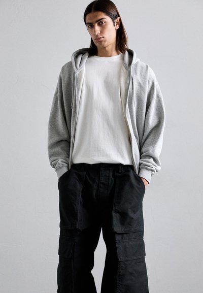 Young person with long hair wearing a grey zip-up hoodie, white t-shirt, and black cargo pants with hands in pockets against a plain background.