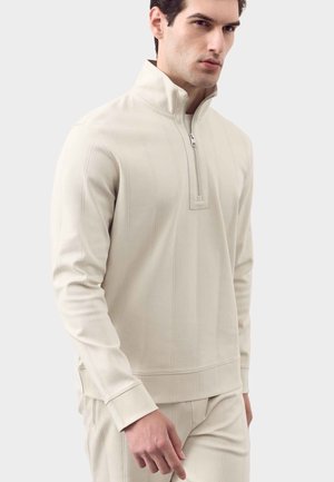 Jonge man draagt een beige trui met een kwart rits en een bijpassende broek, staand tegen een effen lichte achtergrond.