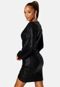Černé šaty s flitry a dlouhými rukávy, přiléhavý střih a texturovaná tkanina. Výrazný lesk od flitrů, délka po stehna, a kulatý výstřih.