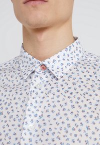 Camicia bianca con un piccolo motivo floreale blu e giallo, caratterizzata da un colletto semplice, chiusura con bottoni anteriori con un bottone rosa e una morbida texture del tessuto.