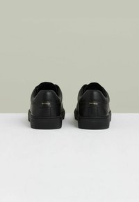 Zapatillas de cuero negro con un diseño elegante, punta redondeada y suela de goma texturizada. Marca dorada notable en la parte trasera.