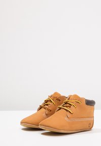 Jasnobrązowe skórzane buciki dziecięce ze żółtymi sznurowadłami i brązowymi akcentami, posiadające miękką, teksturowaną powierzchnię, przeszycia oraz zaokrąglony kształt czubka.