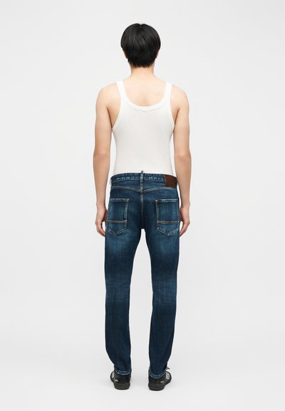 Personne aux cheveux courts noirs portant un débardeur blanc, un jean bleu avec de grandes poches arrière et des chaussures noires, debout de dos.