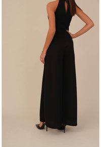 Vrouw in een zwarte mouwloze jumpsuit met wijde pijpen, zwarte hoge hakken, gouden armband en ringen, staand tegen een beige achtergrond.