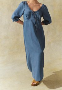 Denim-maksimekko V-pääntiellä, puhvihihat, vyötäröllä kiristysnauha ja sileä pinta vaaleansinisessä sävyssä.