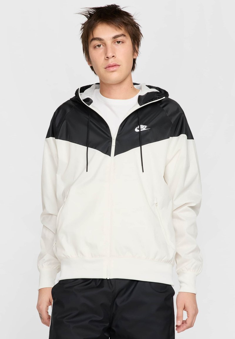 Biela a čierna farba blokovaná bunda so zipsom, kapucňou na šnúrku a elastickými manžetami. Na hrudi sa nachádza logo Nike. Hladká textúra.