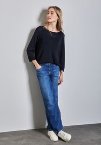 Marineblauwe gebreide trui, met een ronde hals, losse pasvorm, gecombineerd met blauwe wijde jeans en witte sneakers, staande tegen een grijze muur.