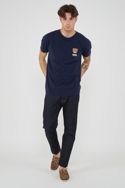 Man with tattoos wearing navy blue teddy bear logo t-shirt, dark jeans, and brown boat shoes, standing against plain light background.