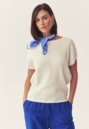 Žena v bílém pleteném topu s krátkým rukávem, modrých kalhotách se šňůrkou v pase, modrém vzorovaném šátku na krk a stříbrných náušnicích, stojící s rukama v kapsách.