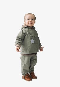 Grüne wasserdichte Jacke mit Fronttasche und Sternen-Detail, kombiniert mit passenden Hosen und braunen Lederschuhen. Weiche, warme Innenausstattung.