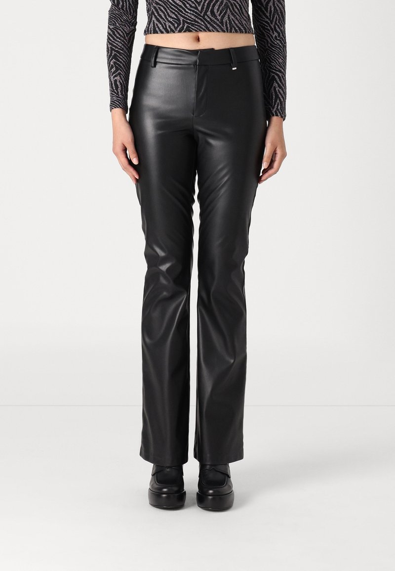 Femme portant un pantalon droit en simili-cuir noir, des chaussures plateformes noires et un crop top à manches longues à motif, debout devant un fond blanc.