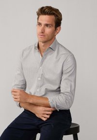 Šedá a bílá kostkovaná košile na knoflíky s dlouhými rukávy, manžetami, v kombinaci s tmavě modrými kalhotami. Má měkký materiál a klasický límeček.