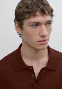 Polo shirt marrone con tessuto testurizzato, una chiusura a tre bottoni e colletto, indossato da un giovane con capelli corti e ricci.