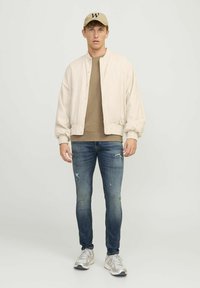 Chaqueta bomber beige, sudadera marrón, jeans azul desgastados y zapatillas gris. La gorra es color beige con un logo "W" negro. Conjunto casual.