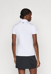 Camisola polo branca com mangas curtas e colarinho, apresentando um logótipo preto nas costas. Combinada com uma saia preta, que mostra uma textura suave.