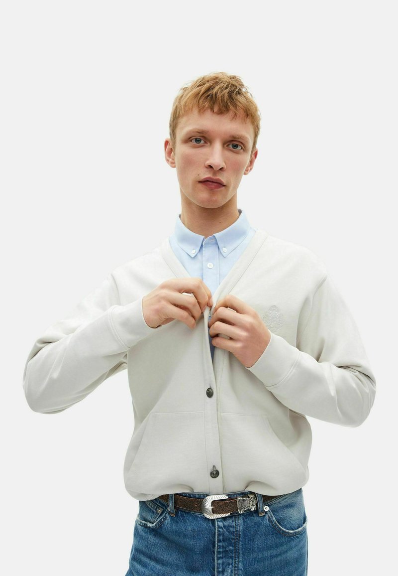 Junger Mann, der einen hellbeigen Cardigan über einem hellblauen Hemd mit Kragen zuknöpft, trägt Jeans und einen braunen Gürtel mit einer silbernen Schnalle, vor weißem Hintergrund.