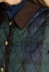 Jachetă verde matlasată cu guler din căptușeală maro, nasturi aurii și un logo brodat. Prezintă un model în carouri diagonale și o țesătură texturată.