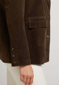 Blazer en suede marron avec une coupe ajustée, poche poitrine simple et fermeture à quatre boutons sur les manches. Texture lisse et design raffiné.