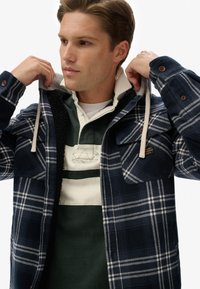 Marineplaidjakke med sherpa-foring, forlommer og knaplukninger. Underneath en grøn og creme-stribet polo skjorte.