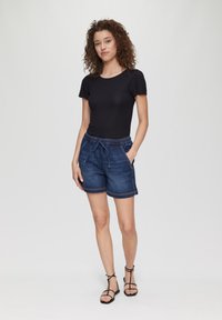Schwarzes T-Shirt mit kurzen Ärmeln, kombiniert mit dunkelblauen Jeansshorts mit elastischem Bund, Kordelzug und Seitentaschen. Schwarze Sandalen mit Riemchen.