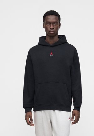 Jonge man met een zwarte hoodie met een rood logo en een witte broek, staand tegen een egale lichte achtergrond.