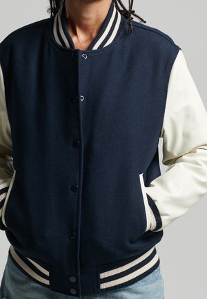 Personne portant une veste universitaire bleu marine et blanche avec des poignets et un col rayés, les mains dans les poches, sur un fond gris uni.