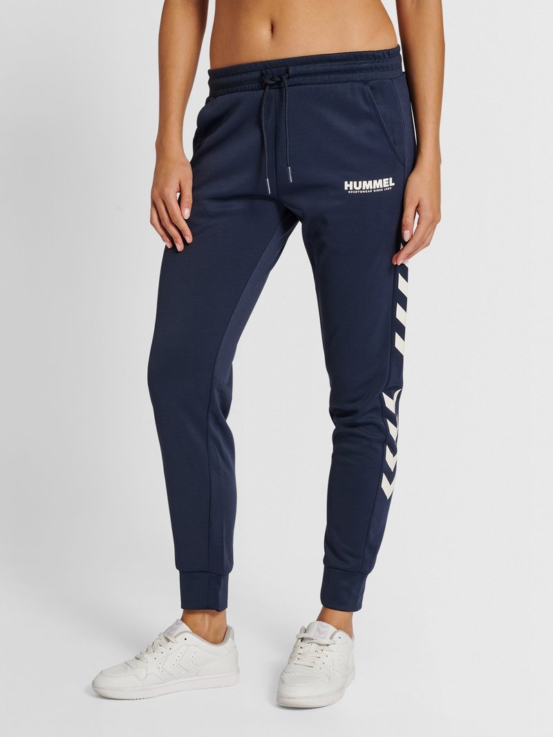 Femme portant un pantalon de survêtement Hummel bleu marine avec des bandes blanches en chevron sur le côté et des baskets blanches, debout devant un fond uni.