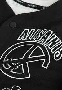 Camisa negra abotonada con logo blanco de AllSaints y símbolo de la paz estilizado en el pecho, sobre una camisa blanca de cuello AllSaints.