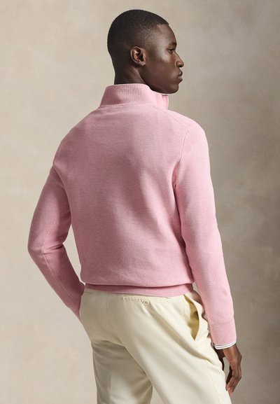 Ροζ πλεκτό πουλόβερ με ψηλό γιακά, ριμπ μανσέτες και μέση. Φορεμένο με ανοιχτό μπεζ παντελόνια, εμφανιζόμενο από πίσω, αναδεικνύοντας την υφή.