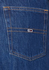 Bolsillo de mezclilla azul oscuro con costuras amarillas, con un pequeño logotipo bordado en rojo, blanco y azul en la solapa.