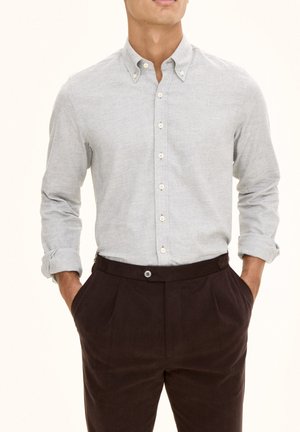Camicia grigia chiara con tessuto testurizzato, colletto classico e maniche arrotolate, abbinata a pantaloni scuri a pieghe marrone.