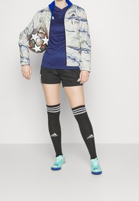 Adidas sportset med en ljus jacka med marmor-mönster, blå skjorta, svarta shorts och strumpor som går till knäna. Blå och grå fotbollsskor.