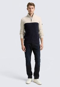 Strickpullover mit cremefarbenem und marineblauem Farbblock-Design, Quarter-Zip-Kragen, gerippten Bündchen und gesprenkelter Textur. Kombiniert mit dunkler Hose.