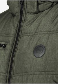 Giacca a vento verde con finitura testurizzata, tasche con zip e una patch rotonda nera sul lato superiore sinistro.