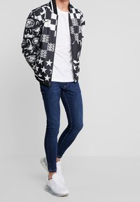 Černá bomber bunda s grafickými vzory, bílými hvězdami a logy, kombinovaná s úzkými tmavými džínami a bílými teniskami.