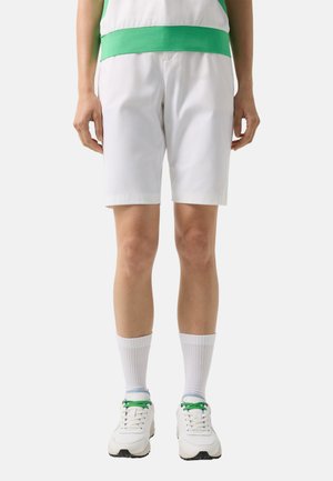 Person iført hvide knælange shorts, hvide sokker og hvide sneakers med grønne snørebånd, stående mod en ensfarvet baggrund.