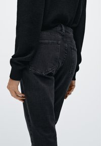 Černé džíny z denimu zobrazené z boku, s vysokým pasem, pěti kapsami a jemnou texturou. Látka má mírné vyblednutí.