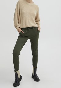 Beige gebreide trui, comfortabele pasvorm. Olijfgroene tapering broek met een trekkoord in de taille. Zwarte laarzen met beige accenten en grove zolen.