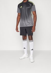 Grå atletisk tröja med gula accenter, svarta shorts, vita strumpor med ränder och vita klackar. Har ett texturerat mönster på tröjan.