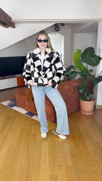 Chaqueta de piel sintética de cuadros en blanco y negro, corte oversized, mangas largas; combinada con jeans de campana de color azul claro y zapatos blancos, en un ambiente interior.