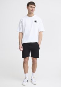 Camiseta blanca de algodón con un gráfico negro en el pecho, combinada con pantalones cortos negros y zapatillas blancas con rayas negras y un logo.