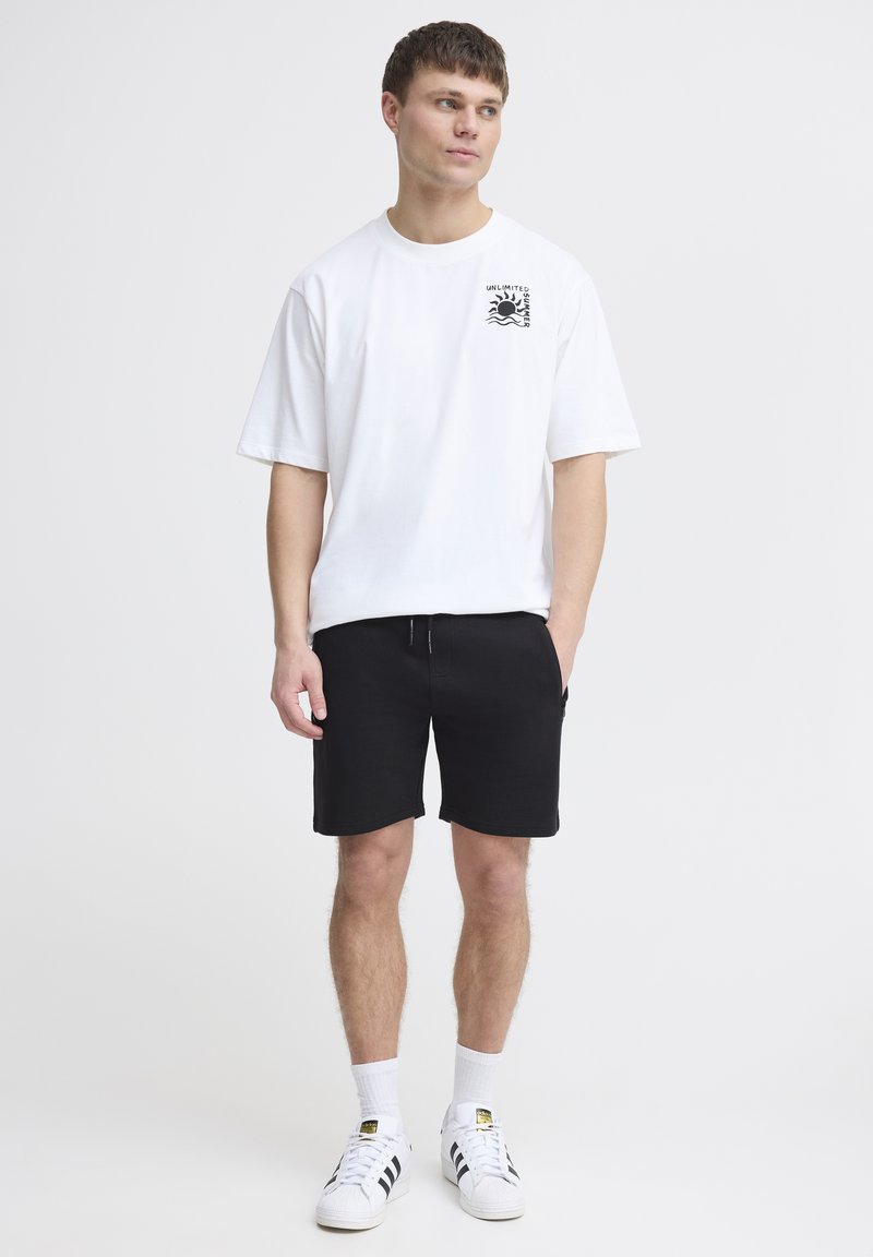 Camiseta blanca de algodón con un gráfico negro en el pecho, combinada con pantalones cortos negros y zapatillas blancas con rayas negras y un logo.
