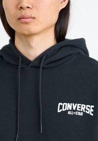 Sudadera negra de tejido suave con un bolsillo canguro frontal, cordones y el logo blanco de "Converse All-Star".