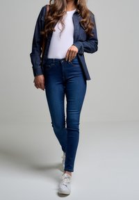 Camisa de mezclilla de botones, camiseta blanca ajustada, jeans ajustados de color azul oscuro y zapatillas blancas. Enfócate en elementos de estilo casual y contemporáneo.