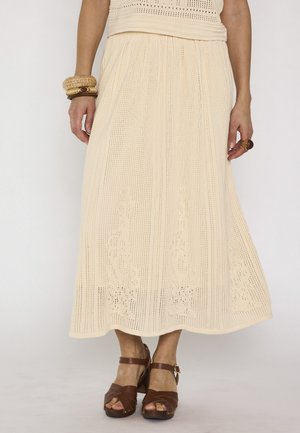 Jupe midi beige en crochet portée avec des sandales marron à talons, des bracelets en bois au poignet gauche et une bague en bois à la main droite.