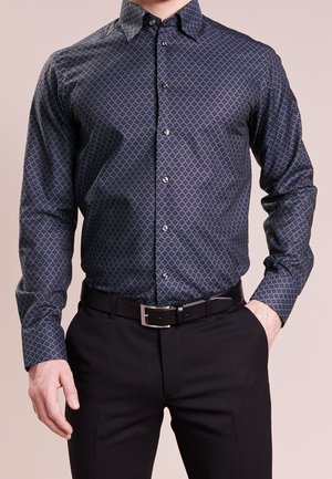 Homme portant une chemise à motifs foncés à manches longues boutonnée et rentrée dans un pantalon noir avec une ceinture noire, main dans la poche.