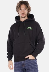 Felpa nera con cappuccio realizzata in un tessuto morbido, dotata di tasca frontale e coulisse. Presenta una scritta verde che recita "PROPAGANDA."