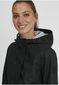 Black waterproof jacket with a drawstring hood, snap buttons, and striped inner lining. Features branded logo patch on the sleeve.