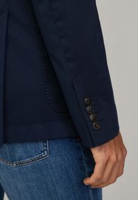 Marineblazer aus strukturiertem Stoff, mit vier dunklen Knöpfen am Ärmel und einer Taschen-Detail, kombiniert mit jeansblauen Jeans.