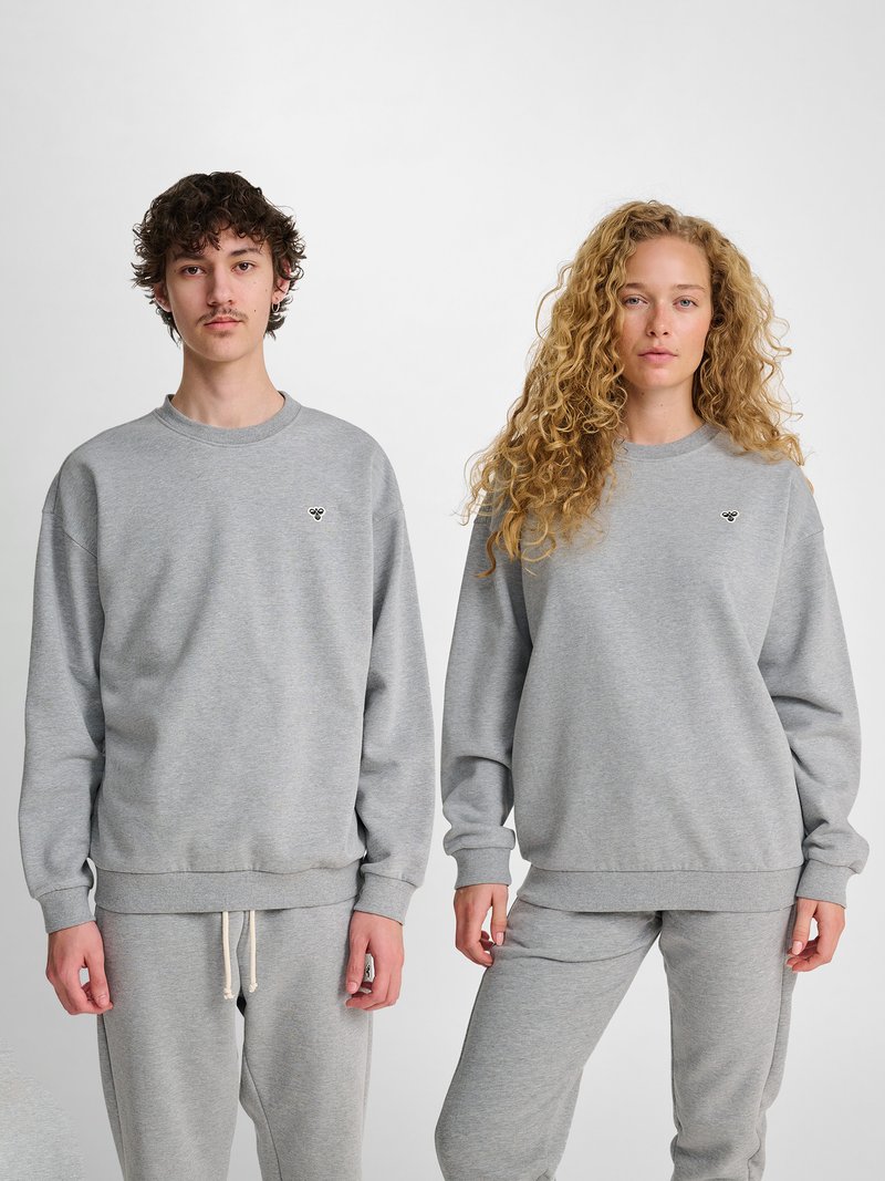 Ung mand og kvinde står side om side iført matchende grå sweatshirts og joggingbukser mod en ensfarvet hvid baggrund.
